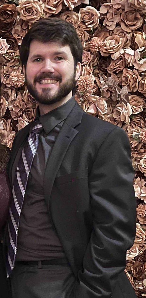 A photo of a bearded person wearing a suit in front of a wall of flowers.